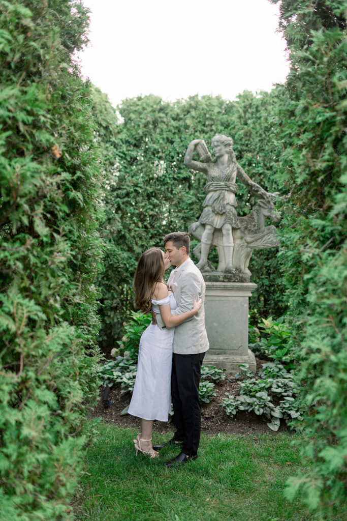 couple kissing by stone statue among dense green bushes